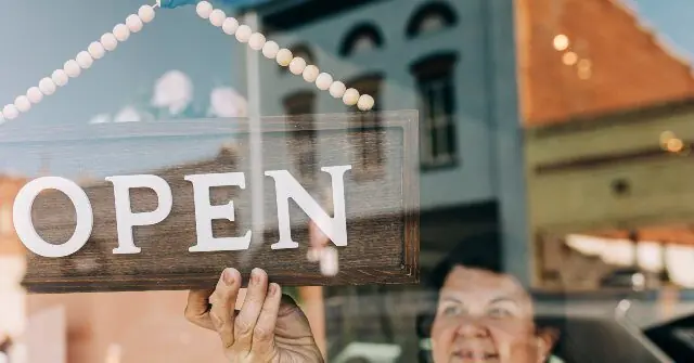 A person is holding up an "OPEN" sign in front of a store window, indicating the business is now open for business.