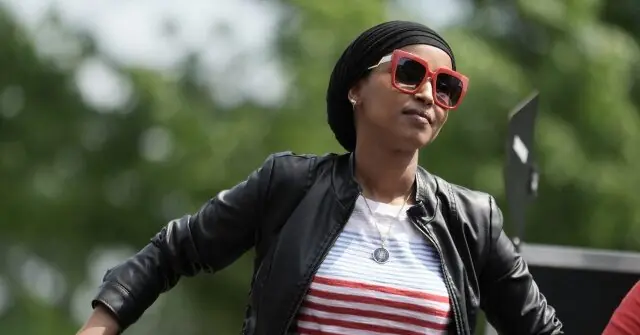 A person wearing a black leather jacket, red-striped shirt, and red-framed sunglasses stands outdoors with a blurred green background.