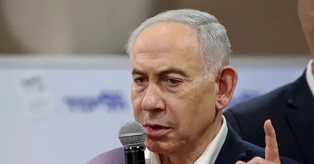 The image shows a man speaking into a microphone, gesturing with his hand, and appears to be in a formal setting, possibly a press conference or public event.