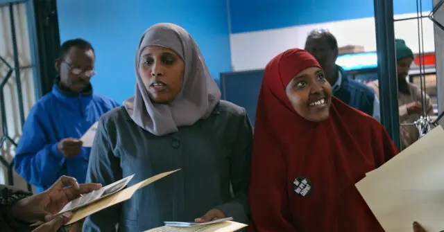 The photo shows a group of people, including two women in hijabs, standing in what appears to be a public or official setting, possibly related to a civic or administrative activity.