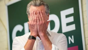 A man covers his face with his hands in front of a sign that reads "CUPDE."