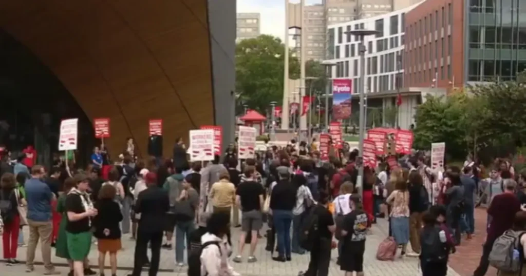 A large group of people gather outdoors, holding signs, possibly participating in a protest or demonstration.