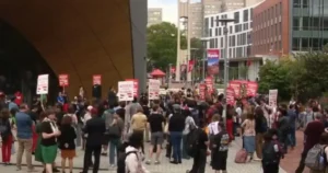 A large group of people gather outdoors, holding signs, possibly participating in a protest or demonstration.
