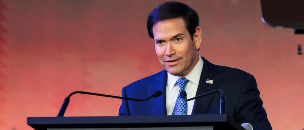 A man in a suit and tie is speaking at a podium with microphones, set against a red background.
