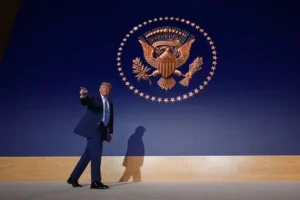 The image shows a person in a suit walking in front of a large projection of the U.S. presidential seal.