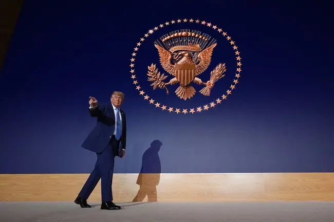 The image shows a person in a suit walking in front of a large projection of the U.S. presidential seal.