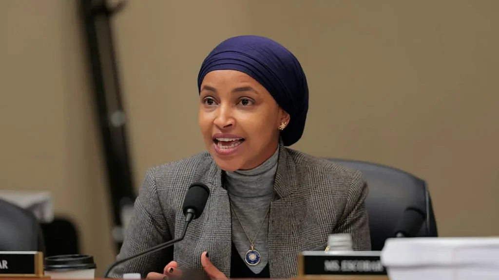 A woman wearing a blue headscarf and a gray blazer is speaking at a formal event, with a microphone in front of her.