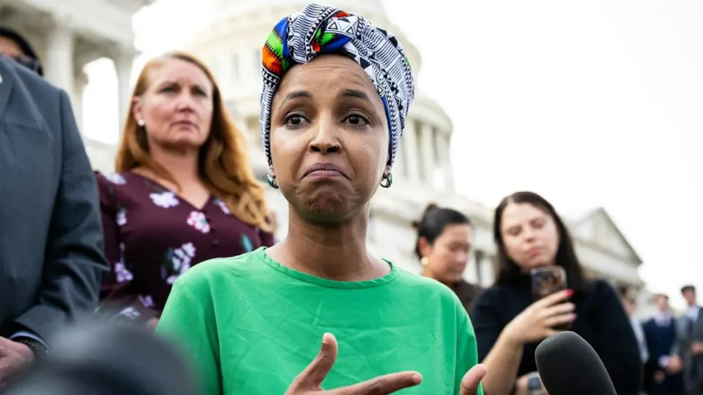 A woman in a green shirt and colorful headscarf speaks to reporters in front of a large building.