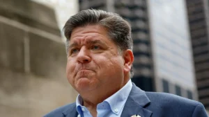 The image shows a man in a suit and tie, standing outdoors with a serious expression, in front of a building.