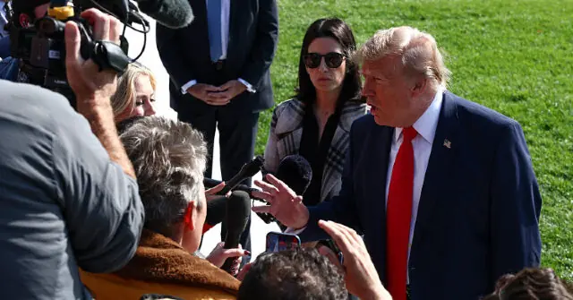 The image shows a man in a suit and red tie speaking to a group of reporters and photographers in an outdoor setting.