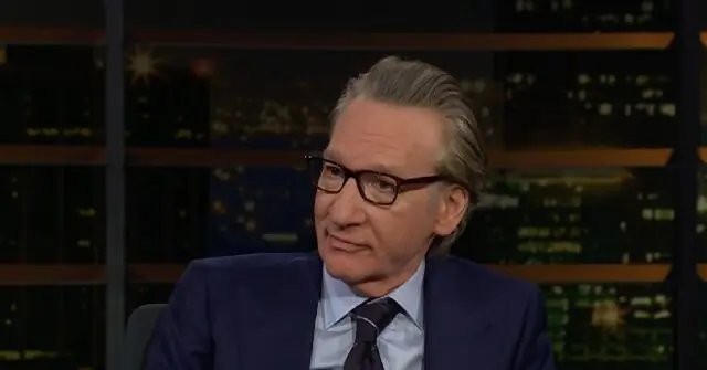 The image shows a man in a suit and tie, wearing glasses, seated in front of a backdrop featuring a cityscape at night.