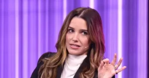The image shows a woman speaking on a stage with a purple background, gesturing with her hand.