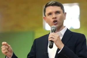 A man in a suit is speaking into a microphone with a fist raised, suggesting a passionate or motivational speech.