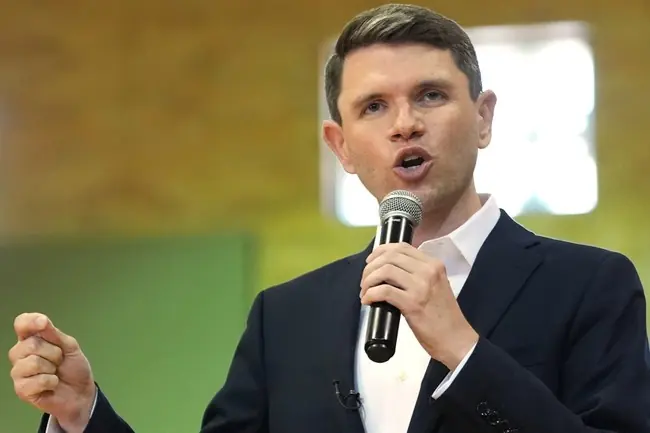 A man in a suit is speaking into a microphone with a fist raised, suggesting a passionate or motivational speech.