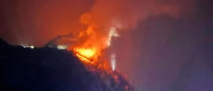 A large fire is burning on a hillside at night, with intense flames and smoke billowing into the sky.