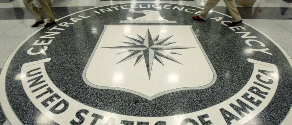 The image shows the logo of the Central Intelligence Agency (CIA) on the floor, with individuals walking past it.