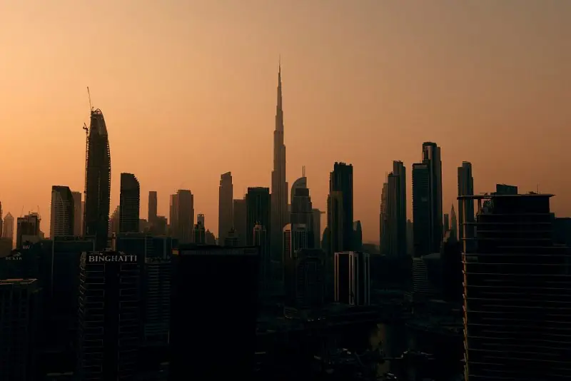 The image captures the silhouette of Dubai's iconic skyline at sunset, showcasing the Burj Khalifa and other modern skyscrapers against a warm, orange-hued sky.
