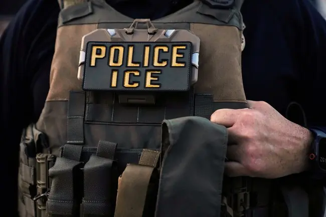 The image shows a law enforcement officer wearing a vest with a "POLICE ICE" patch, indicating their affiliation with Immigration and Customs Enforcement (ICE).