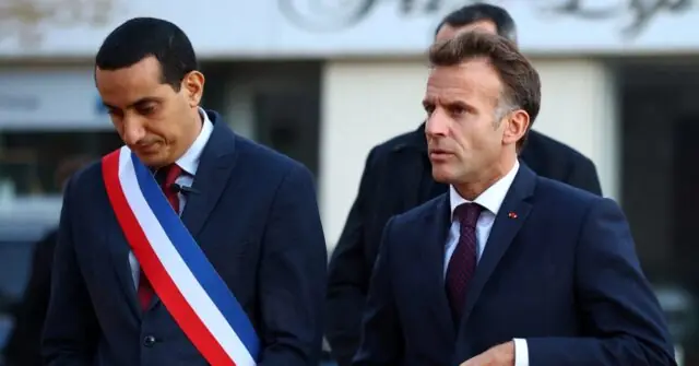 The image shows two men in formal attire, one wearing a red, white, and blue sash, walking together in what appears to be a public or official setting.