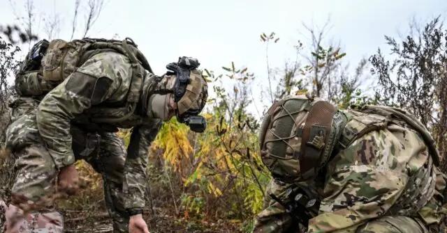Two soldiers in camouflage uniforms are seen in a forested area, with one bending down and the other sitting, possibly engaged in a training or reconnaissance activity.