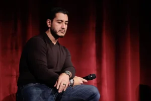 A man is seated on stage, holding a microphone, with a red curtain in the background.