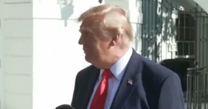 The image shows a man in a suit and red tie standing outside a building with a white facade and ornate metalwork.