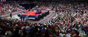 A large crowd of people wearing red hats and patriotic attire is gathered in a stadium to watch a political rally.