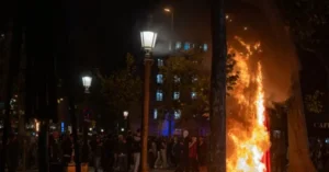 The photo depicts a nighttime scene with a crowd gathered around a large fire, illuminated by streetlights and the glow of the flames.