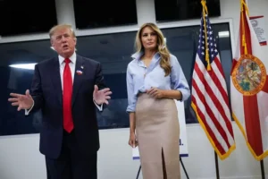 The image shows a man and a woman standing in front of American and Florida state flags, with the man gesturing as if speaking.