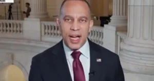 The image shows a man in a suit and tie speaking in front of a backdrop featuring classical architecture, likely a government building.