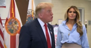 The image shows a man in a suit and a woman in a light blue blouse standing in front of flags, possibly at a public event or press conference.