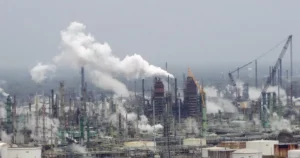 The image shows a large industrial facility with numerous smokestacks emitting plumes of smoke, indicating ongoing industrial activity and potential environmental impact.