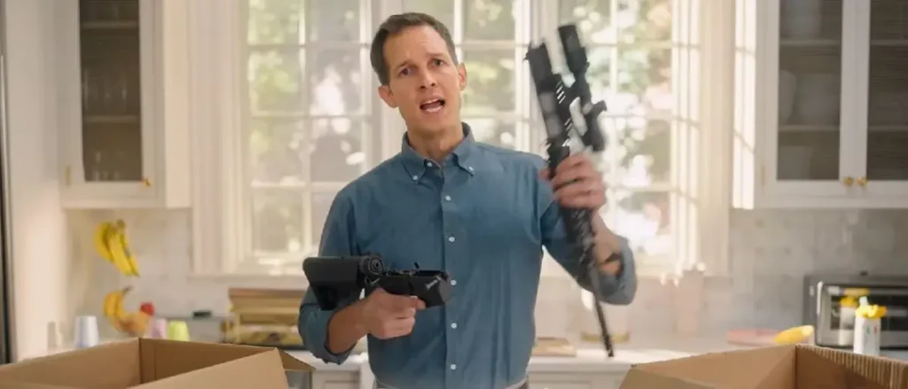 A man in a kitchen holds a paintball gun and a tablet, seemingly reacting to something unexpected.