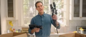 A man in a kitchen holds a paintball gun and a tablet, seemingly reacting to something unexpected.
