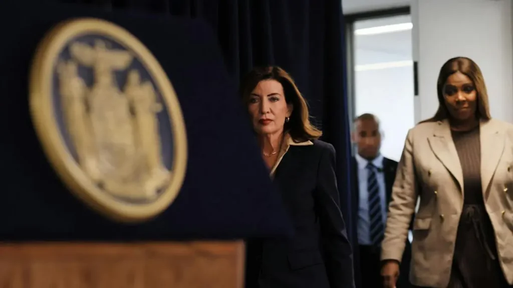 A woman in a suit walks in front of a backdrop featuring a seal, with another woman and a man following behind.
