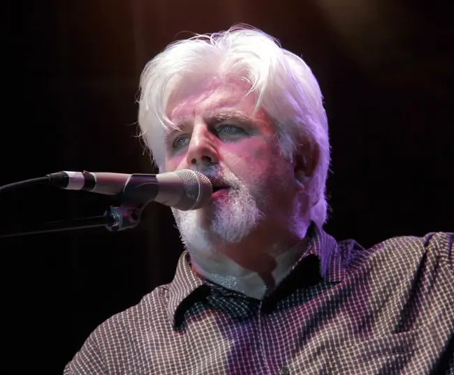 The image shows a person with white hair and a beard, wearing a checkered shirt and a bow tie, singing into a microphone on stage.