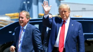 The image shows a man in a blue suit and red tie waving, accompanied by another man in a blue suit and pink tie, as they appear to be disembarking from an aircraft.