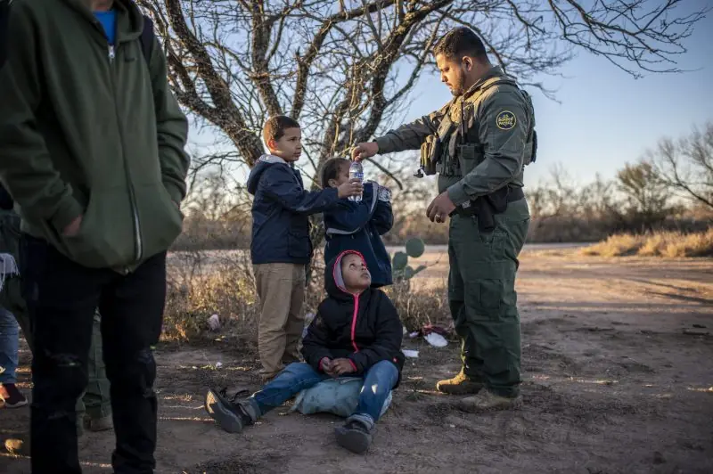A Border Patrol agent offers water to a child while other individuals observe in a rural setting.