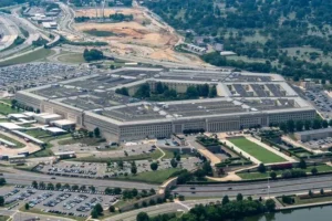The image shows an aerial view of the Pentagon, a large five-sided building located in Washington, D.C., surrounded by roads, parking lots, and greenery.