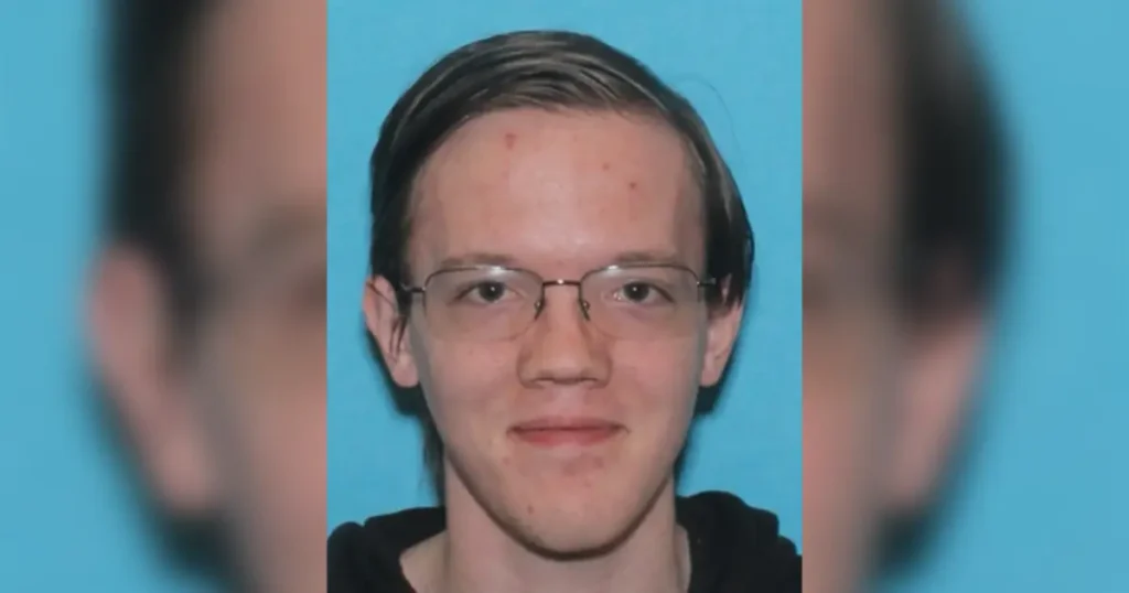 The image shows a person with short hair and glasses, wearing a black top, against a blue background.