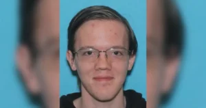 The image shows a person with short hair and glasses, wearing a black top, against a blue background.