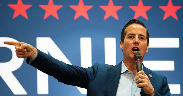 A man in a suit is speaking into a microphone while pointing to the side, with a backdrop featuring red stars and the letters "RNE."