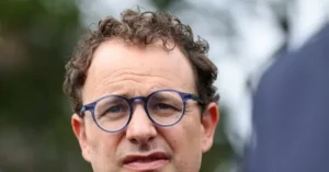 The image shows a close-up of a person wearing glasses, with a blurred background that suggests an outdoor setting.