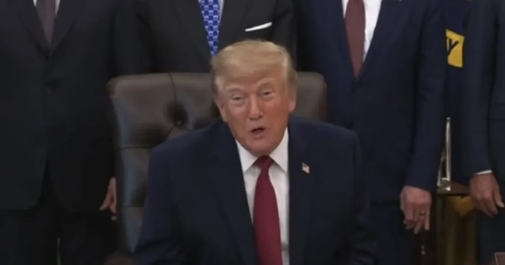 The image shows a man in a suit seated in a formal setting, with other individuals in suits standing behind him.