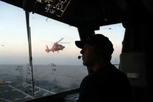 A person is seen inside a cockpit, observing a helicopter flying over the ocean.