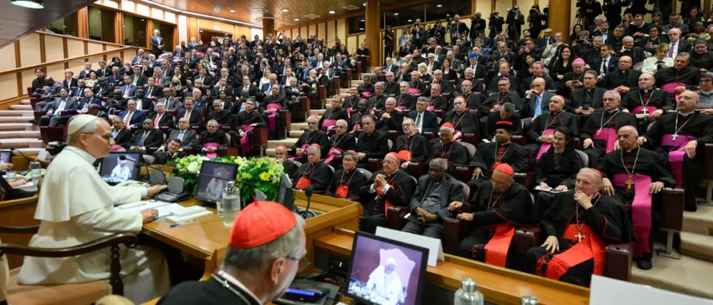 The image depicts a formal gathering in a large hall, with a prominent figure in white addressing a group of clergy and attendees, who are attentively listening and taking notes.