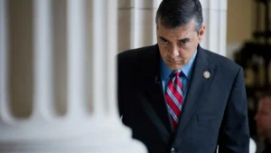 The image shows a man in a suit and tie, standing in a formal setting with columns in the background.