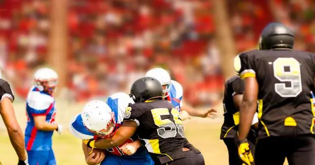 A football player in a blue and white uniform is tackled by a player in a black uniform during a game.