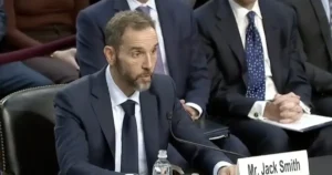 A man in a suit and tie is seated at a table with a microphone, appearing to testify or speak at a formal hearing.