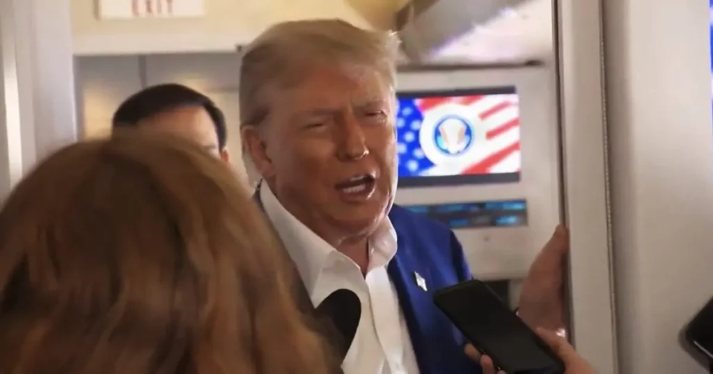 The image shows a man in a blue suit and white shirt speaking to reporters, with a television screen in the background displaying an American flag and a logo.